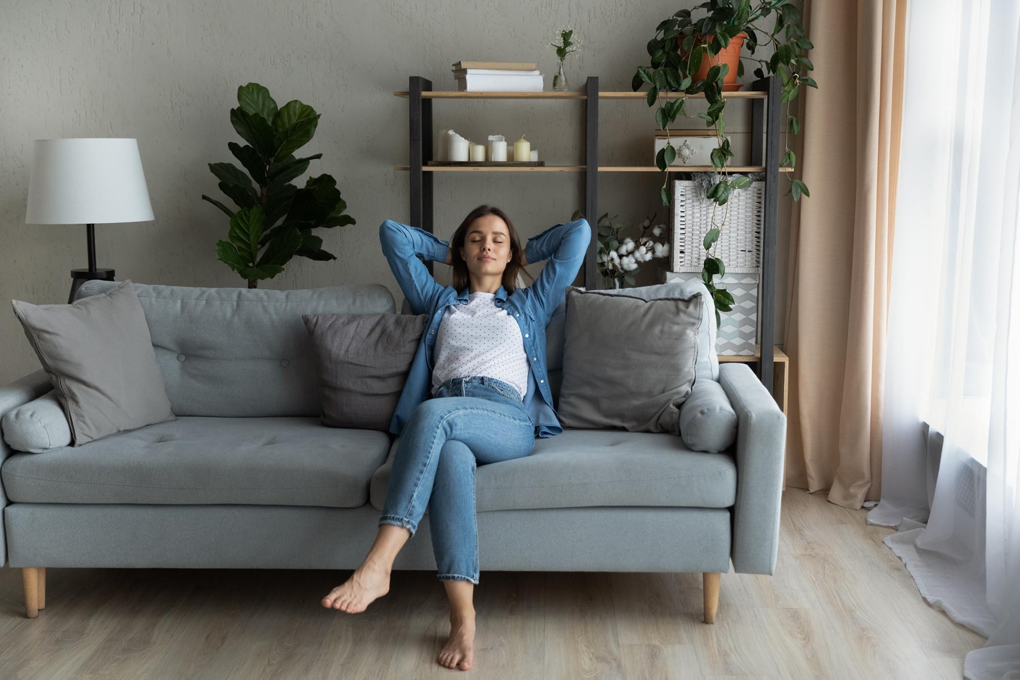 Serene young woman relaxing on comfy sofa with hands behind head, breath fresh conditioned air inside of modern light living room.