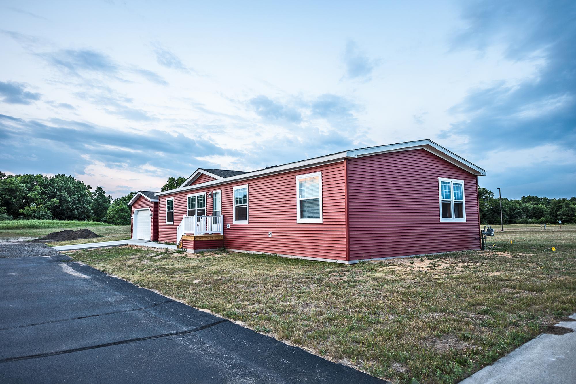 Red manufactured home with one stall garage sitting on a rural tree covered lot